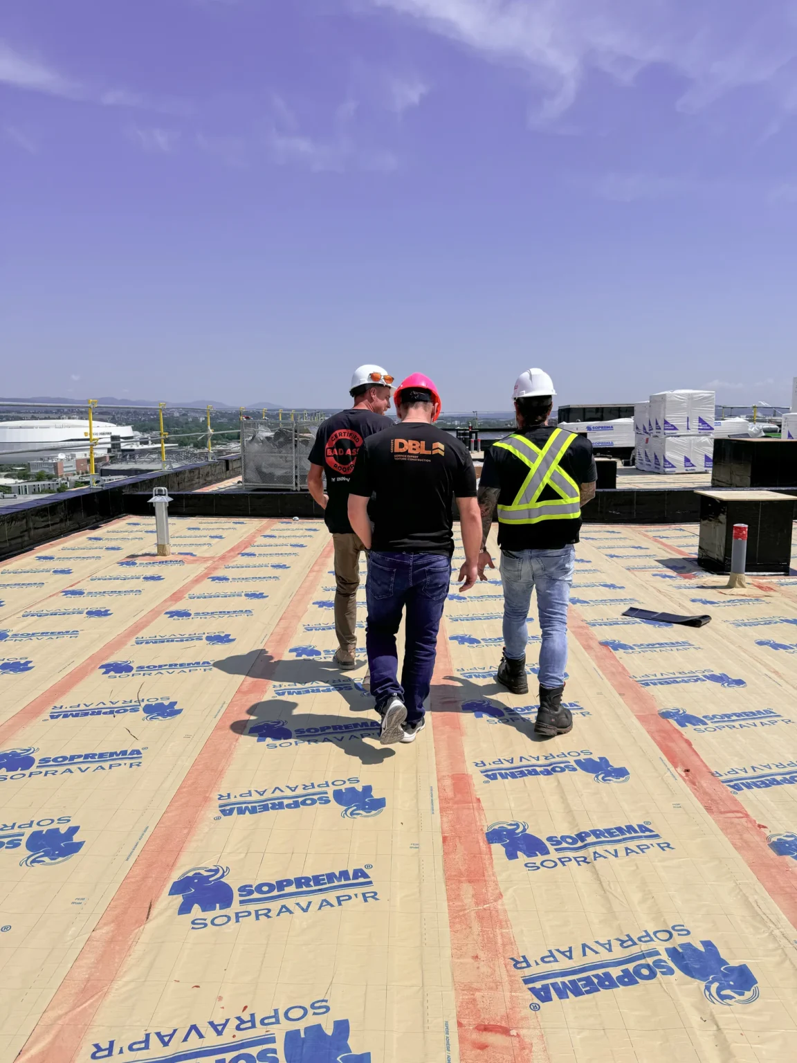 Trois travailleurs sur un toit plat avec membrane Soprema, portant des casques et vêtements de chantier Groupe DBL.