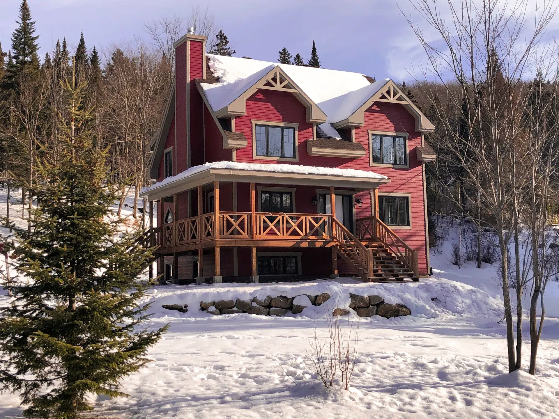 Chalet rouge en bois avec toit enneigé, balcon en bois et forêt en arrière-plan en hiver.